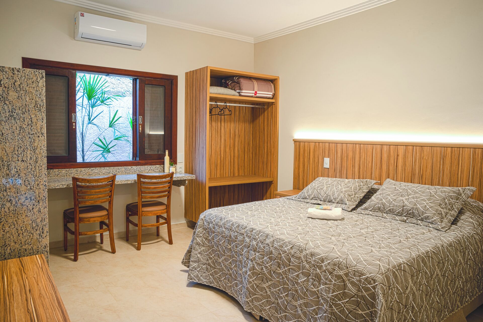 Un dormitorio con cama, armario de madera, escritorio con dos sillas y ventana con vistas a plantas verdes.