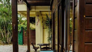 A covered outdoor seating area with wooden tables and chairs, a gravel path, and a green fence in the background.