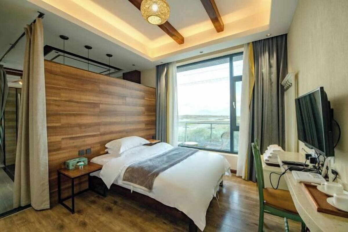 A hotel room with a bed, bedside table, TV, and a large window offering a view.