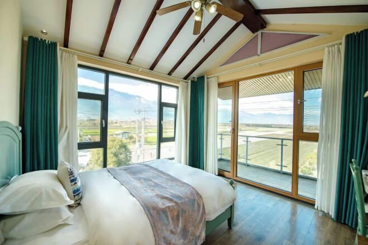 A bedroom with a large bed, a view of the outdoors through a sliding glass door, and a ceiling fan.