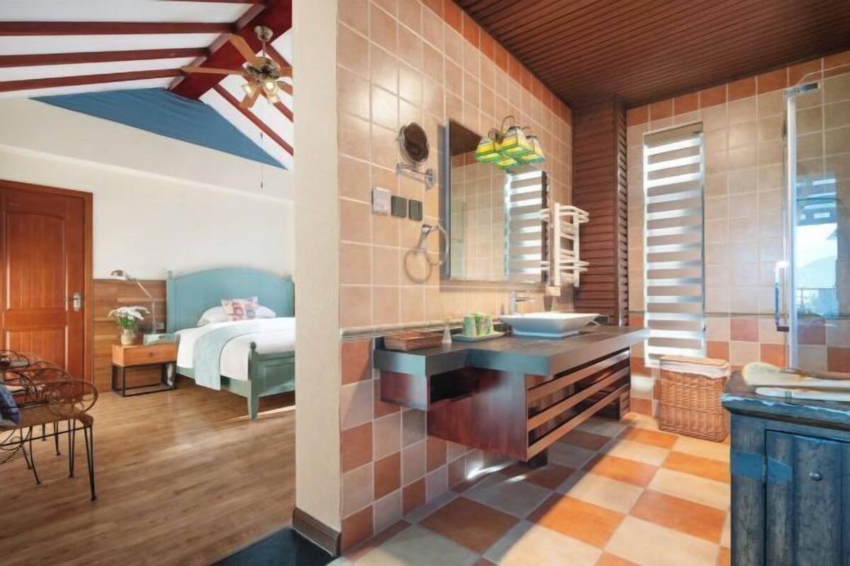 A bedroom with a bed, bedside table, and a chair. There is a bathroom with a sink, mirror, and towel rack. The room has wooden flooring and a sloped ceiling with exposed beams.
