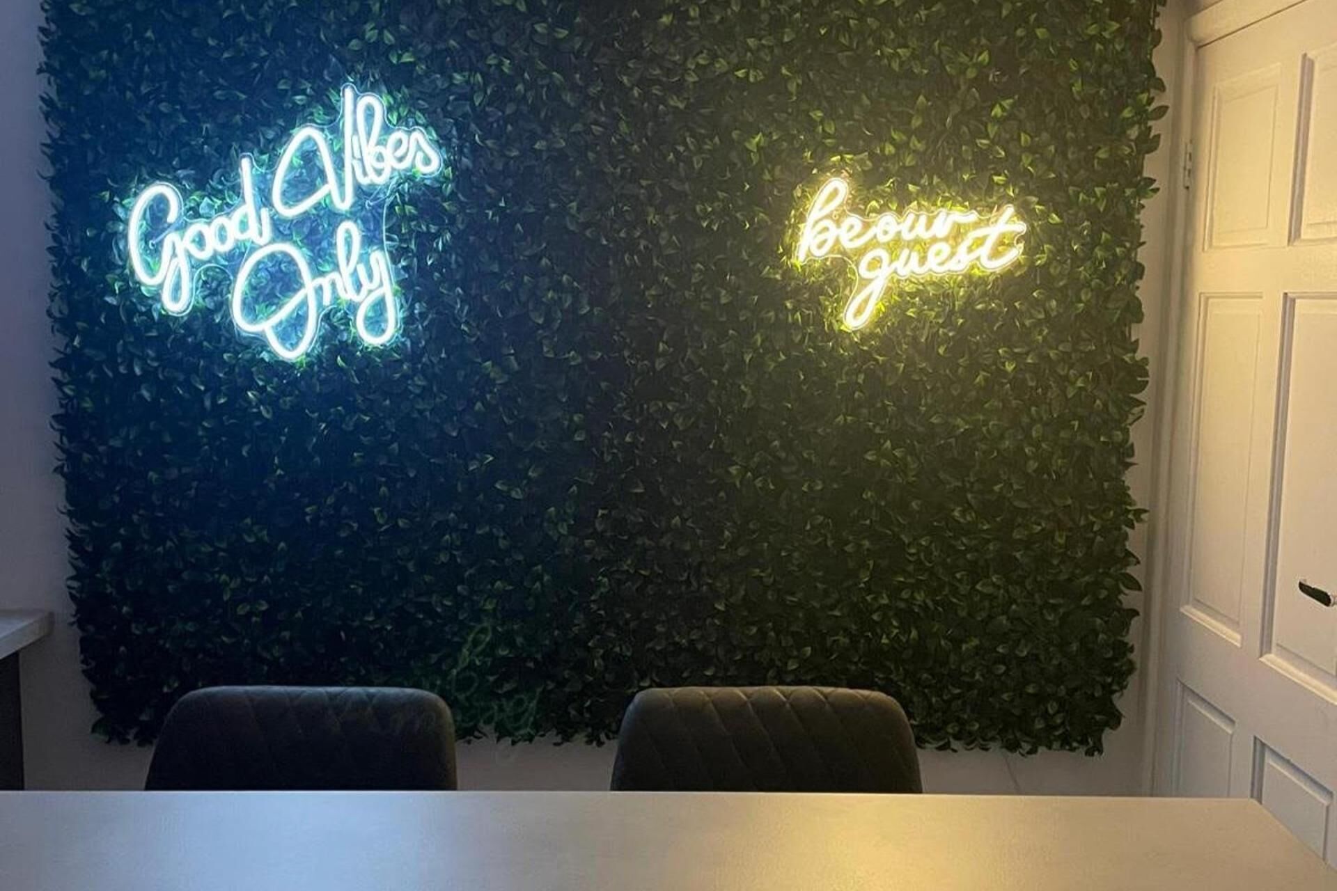 Une pièce dont un mur vert foncé est recouvert de feuillage, et où l’on peut voir deux enseignes lumineuses néon avec les inscriptions « Good Vibes Only » et « Be Our Guest ».