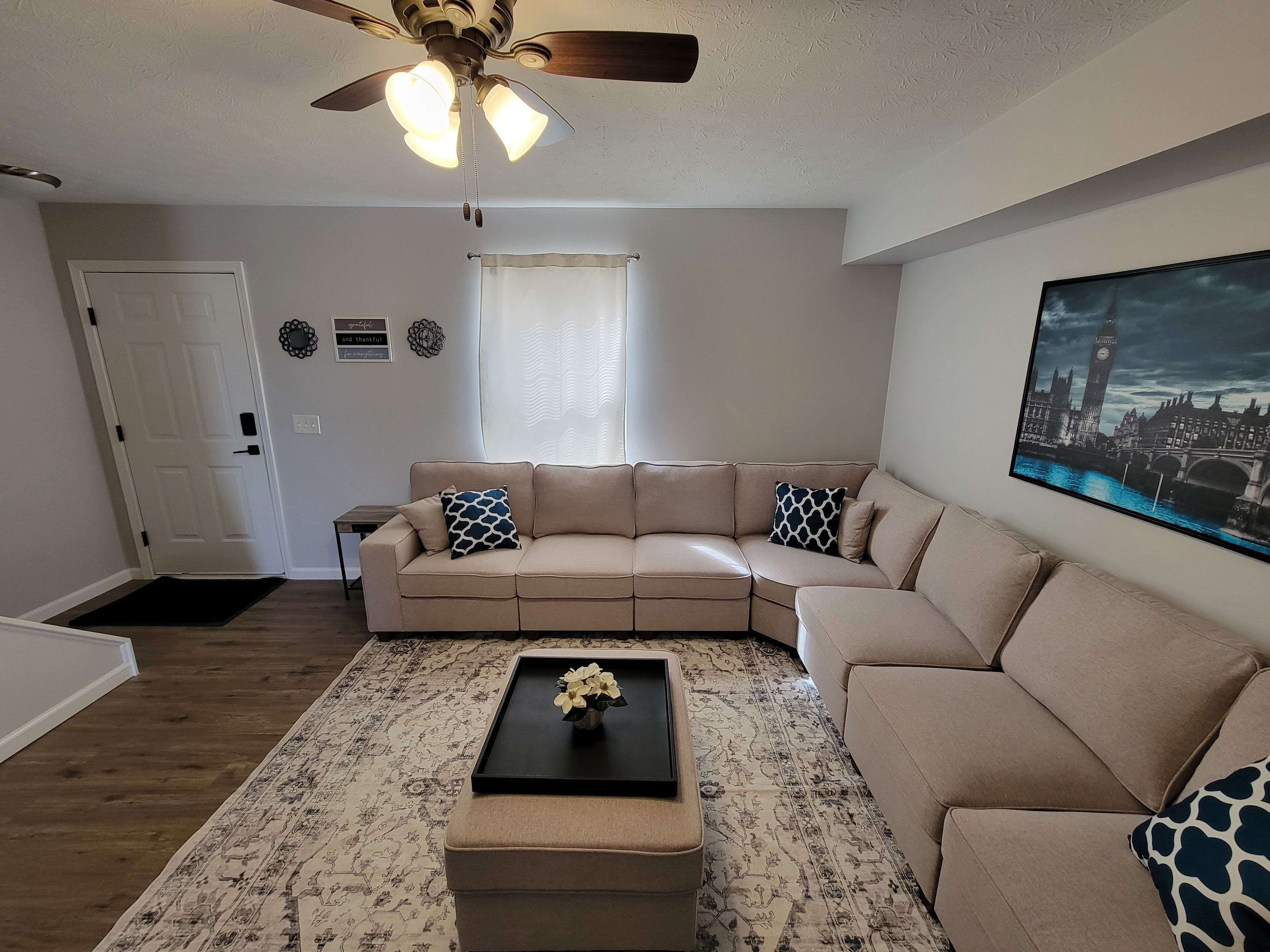 A living room with a beige sectional sofa, a black coffee table with a vase, a patterned rug, a ceiling fan, and a framed cityscape picture on the wall.