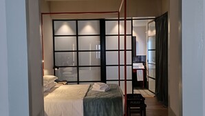 A modern bedroom with a four-poster bed, a large window with blinds, a bedside table, and a view of the bathroom.