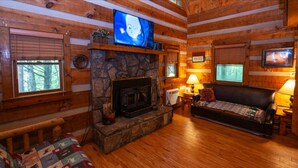 Ein Holzblockhaus-Interieur mit Kamin aus Stein, einem darüber montierten Fernseher, einer Couch und einem Fenster mit Jalousien.