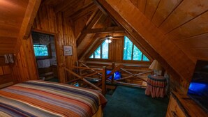 Ein gemütliches Schlafzimmer unter dem Dach mit einem Bett, einem kleinen Hocker, einem Fernseher und einem Fenster mit Blick auf Bäume.