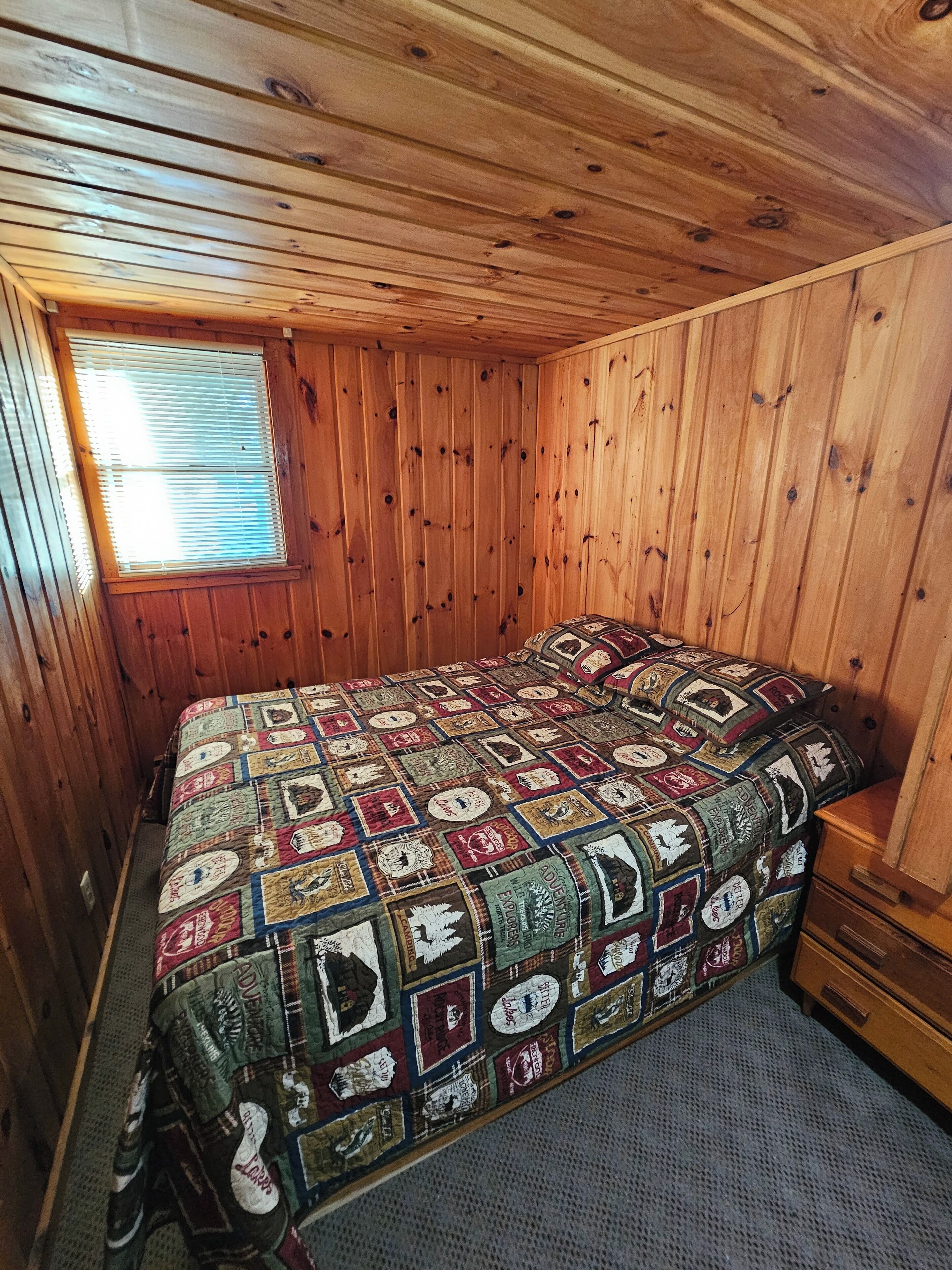 Een houten kamer met een geborduurde spreideken, een raam met jaloezieën en een houten dressoir.