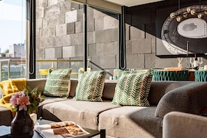 A modern living room with a sofa, patterned cushions, a coffee table with a vase and magazine, and a large window with a city view.