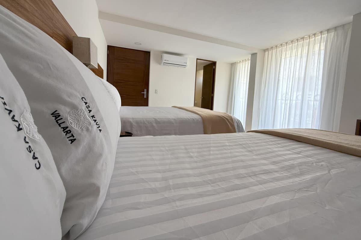 A hotel room with two beds, a wooden headboard, a window with sheer curtains, and a door leading to another room.