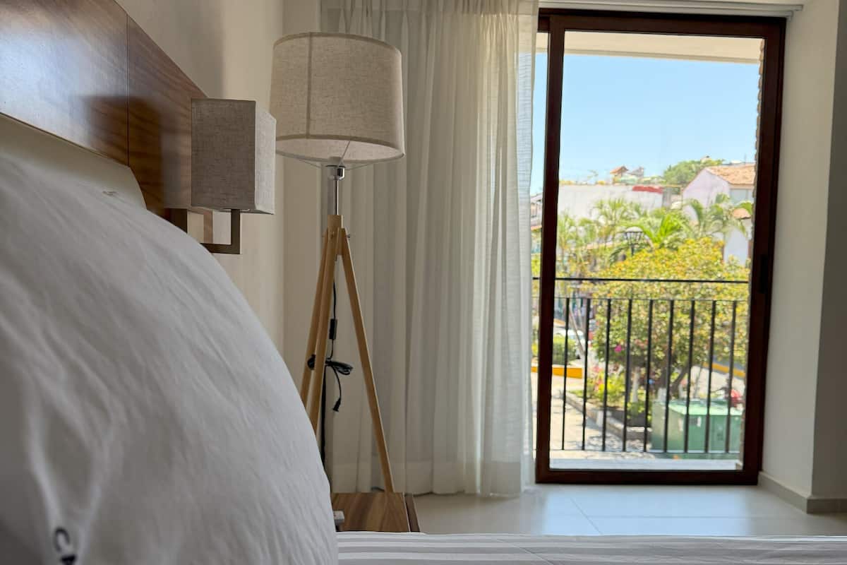 A hotel room with a bed, a floor lamp, a view of a balcony with a railing, and a door leading outside.