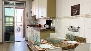 A kitchen with white cabinets, a granite countertop, and a dining area with white chairs and a glass bowl. - Ático con piscina - Guarapari (Guarapari)