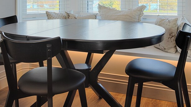 A dining area with a round table and four chairs, large windows with blinds, and a hanging light fixture.