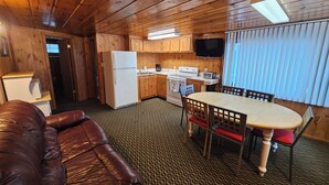 A compact kitchen with wooden cabinets, a refrigerator, a stove, and a microwave. There is a dining table with chairs and a leather couch.