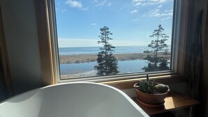 A room with a large window overlooking a beach and trees, a bathtub, and a potted plant on a wooden table. - Lawrencetown Beach House with Hot Tub & Sauna (Head of Chezzetcook)