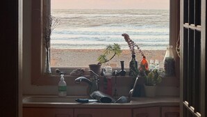 A window with a view of the beach, a sink with various items, and a shelf with decorative objects. - Lawrencetown Beach House with Hot Tub & Sauna (Head of Chezzetcook)