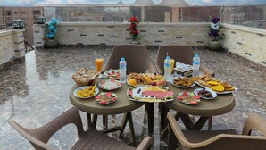 A rooftop dining area with a variety of food and drinks on tables, surrounded by wicker chairs and a view of buildings in the background.
