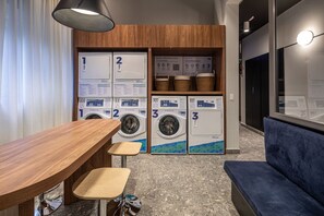 A laundry room with three washing machines, a wooden table, two stools, and a blue sofa. - AD330 (São Paulo)