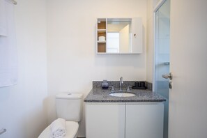 A bathroom with a white toilet, a granite countertop sink, a mirror, and a glass shower enclosure. - AD330 (São Paulo)