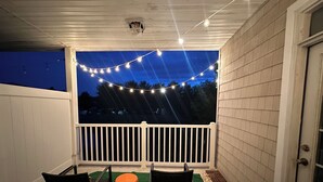A covered patio with a white railing, two chairs, a round orange ottoman, and string lights. - Prime Location In Lewes-  3bed/2bath Great space and game room. (Lewes)