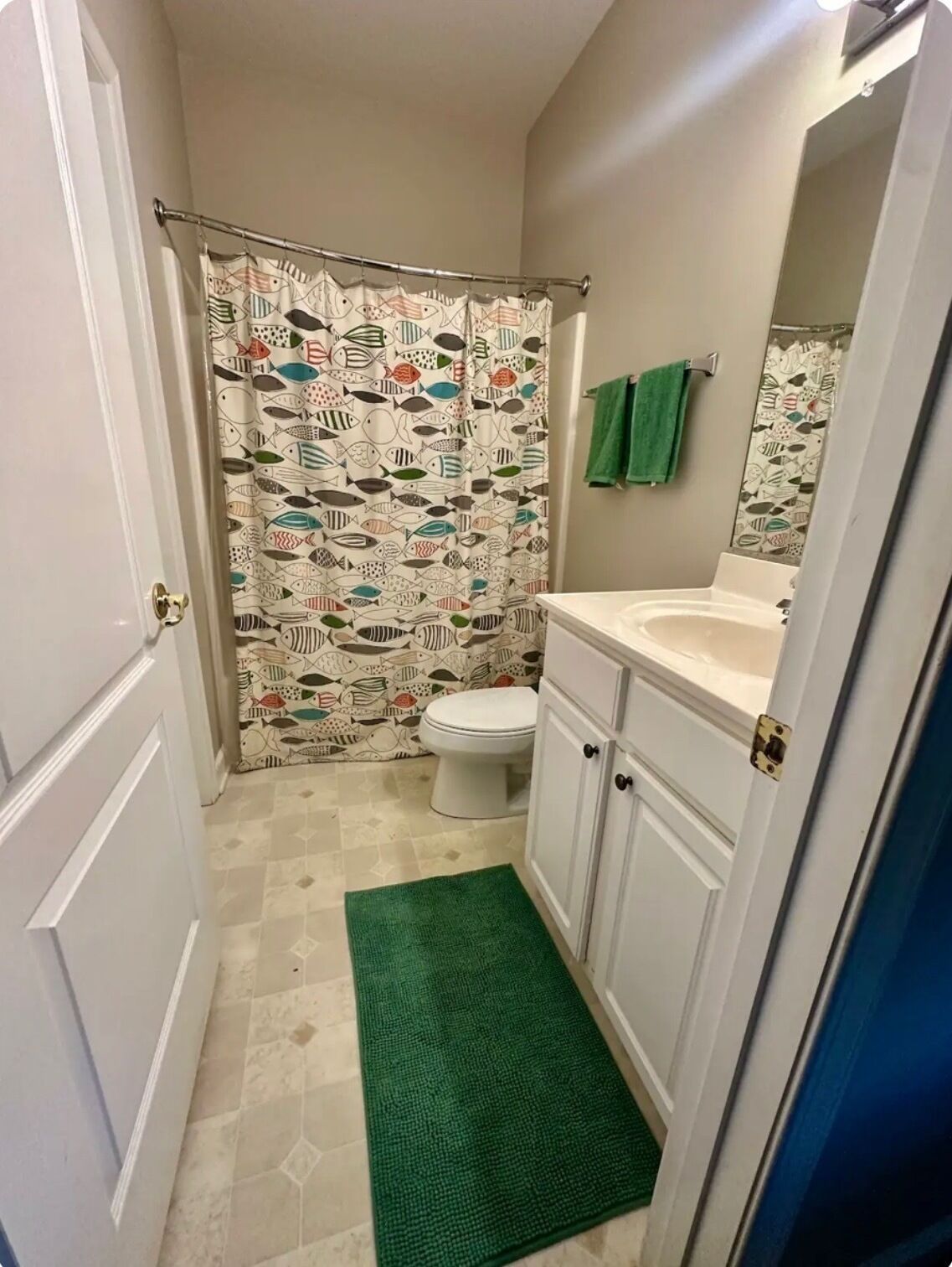 A small bathroom with a shower curtain featuring a fish pattern, a toilet, a sink, and a green rug.