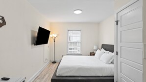 A bedroom with a bed, a TV mounted on the wall, a bedside table, a lamp, and a window with blinds. - Cozy Urban Getaway - 3BR 1.5BA - 7 Mins to Downtown CLT (Charlotte)
