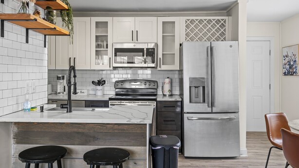 A modern kitchen with white cabinets, stainless steel appliances, a wooden island with stools, and a trash bin. - Cozy Urban Getaway - 3BR 1.5BA - 7 Mins to Downtown CLT (Charlotte)