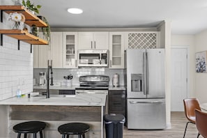 A modern kitchen with white cabinets, stainless steel appliances, a wooden island with stools, and a trash bin. - Cozy Urban Getaway - 3BR 1.5BA - 7 Mins to Downtown CLT (Charlotte)