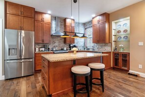 Une cuisine moderne avec des meubles en bois, un réfrigérateur en acier inoxydable, un îlot central doté d’un comptoir en granit et de tabourets, et un pare-feu dont la texture rappelle celle de la pierre.