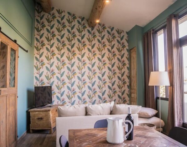 A cozy room with a patterned wallpaper, a wooden ceiling, a bed with pillows, a nightstand, a lamp, and a bottle of wine. - Charming, quiet T1 (Saint-Andiol)