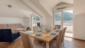 A dining area with a wooden table set for four, a sofa, and a view of a mountainous landscape through large windows. - Stylish Budva Apt withPanoramic View-2Bedroom Apt (Budva)