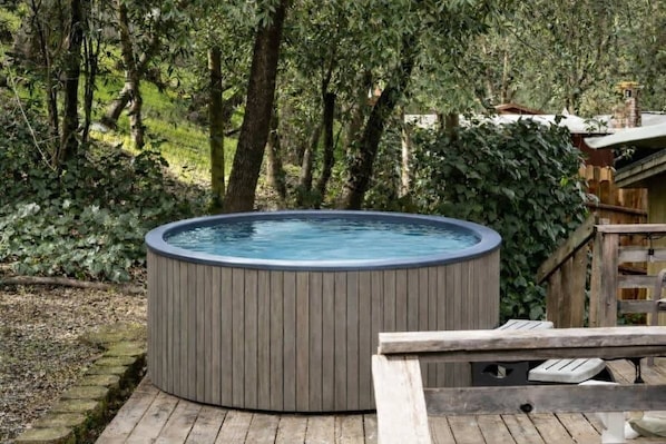 A hot tub in a wooded area with a wooden deck and a small building in the background.