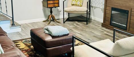 Un salón con un sofá de cuero marrón, un sillón blanco, un tapiz estampado, una mesa de centro de madera, una mesita con una lámpara, un sillón con un cojín amarillo y una chimenea.
