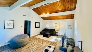 A spacious living room with a high wooden ceiling, a black sofa, a bean bag, and a staircase leading to another level.