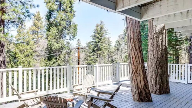 A wooden deck with Adirondack chairs and a large tree trunk, surrounded by a white railing and dense forest in the background.