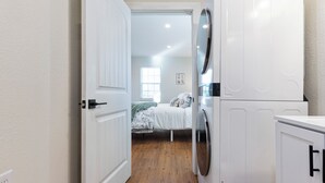 Uma lavanderia com máquina de lavar e secadora, uma cama com cobertor verde e uma janela com persianas.