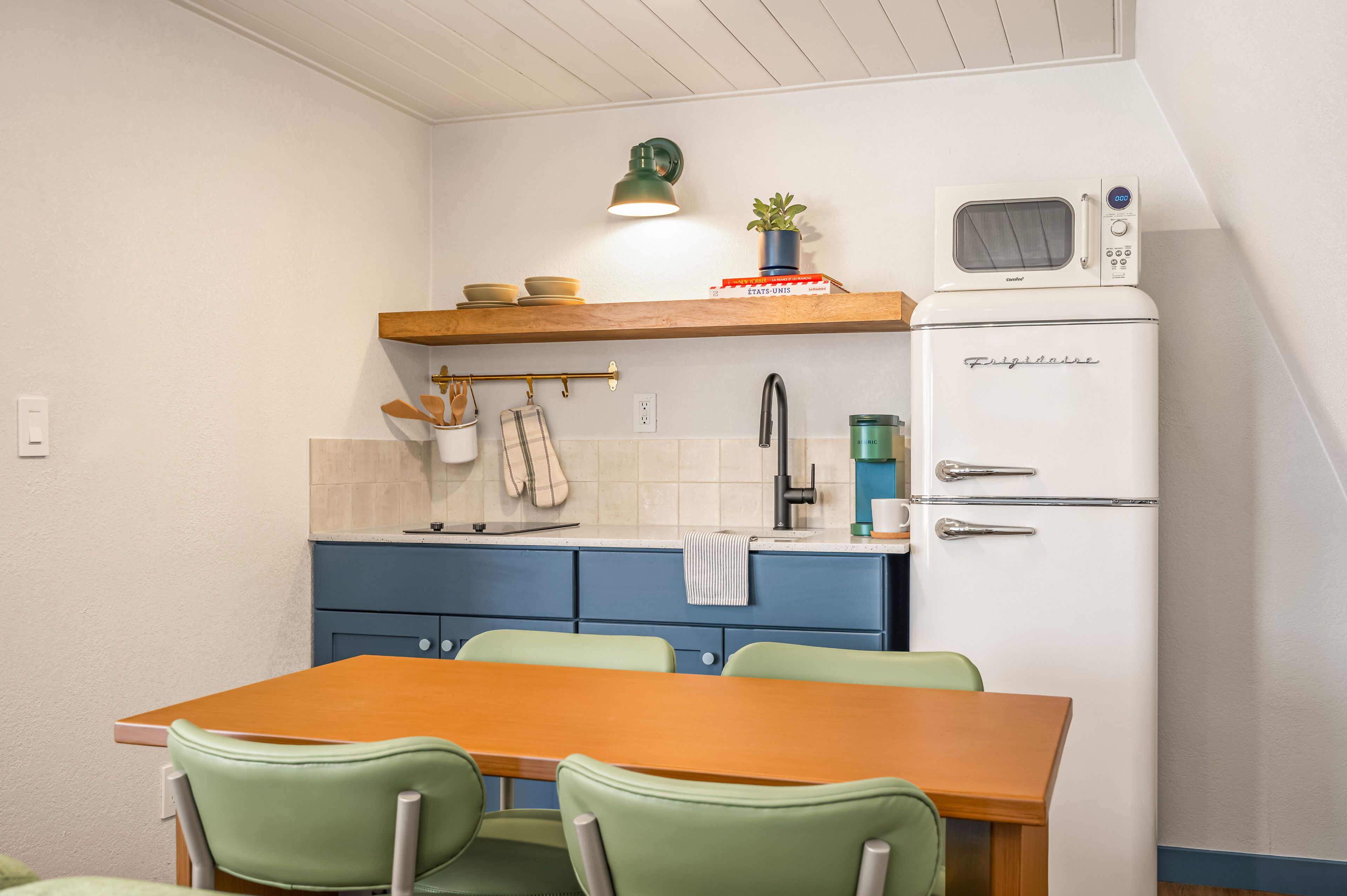 A kitchen with a wooden table and green chairs, a white refrigerator, and a shelf with dishes and a plant.