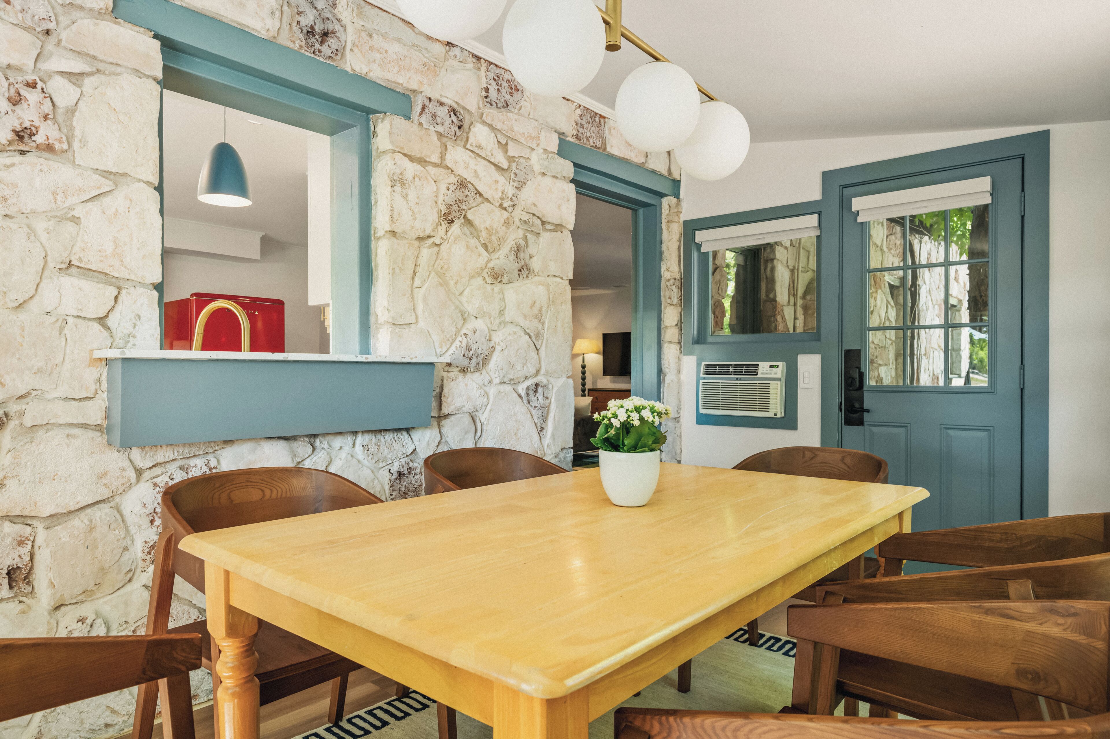 A dining area with a stone wall, wooden table, chairs, a potted plant, and a red appliance in the background.