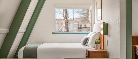 Un dormitorio con cama, mesita de noche, lámpara y ventana con vistas a árboles y edificios.