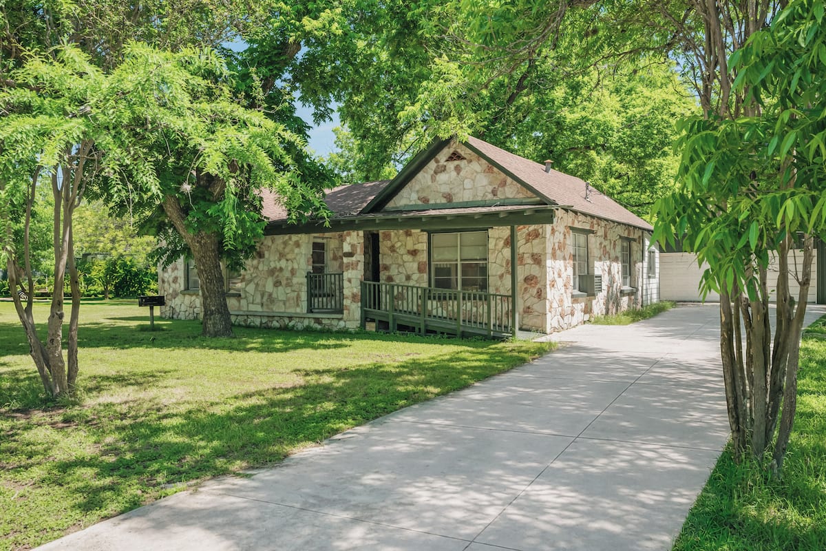 A small stone building with a porch, surrounded by trees and a sidewalk.