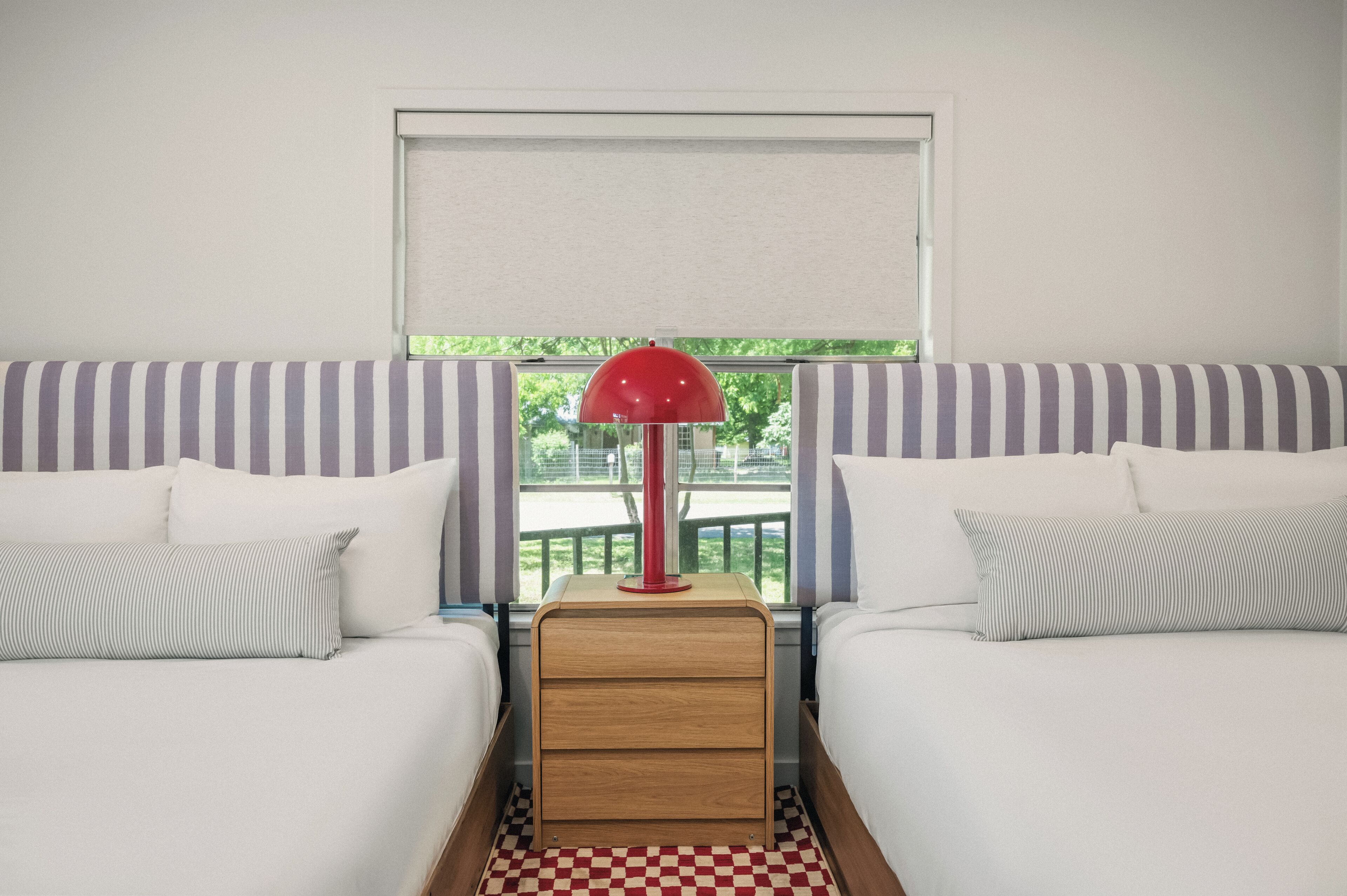 A room with two beds, a window with a view, and a red lamp on a wooden nightstand.