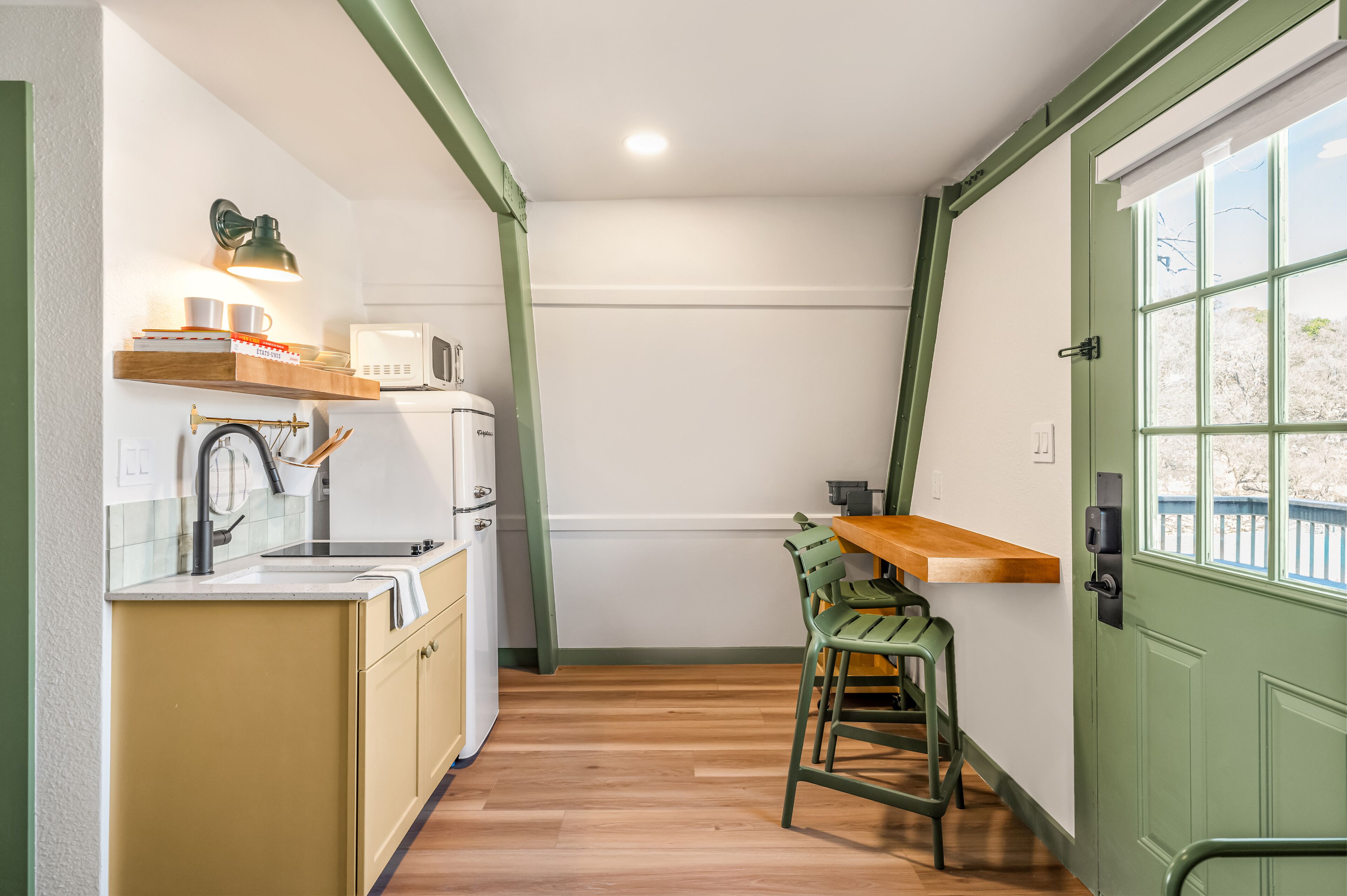 Cocina compacta con microondas, pileta y encimera de madera. Una silla verde está colocada junto a la encimera, y se ve una ventana con vista a los árboles.