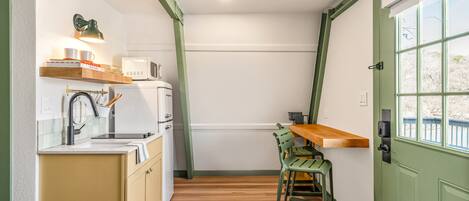Cocina compacta con microondas, pileta y encimera de madera. Una silla verde está colocada junto a la encimera, y se ve una ventana con vista a los árboles.