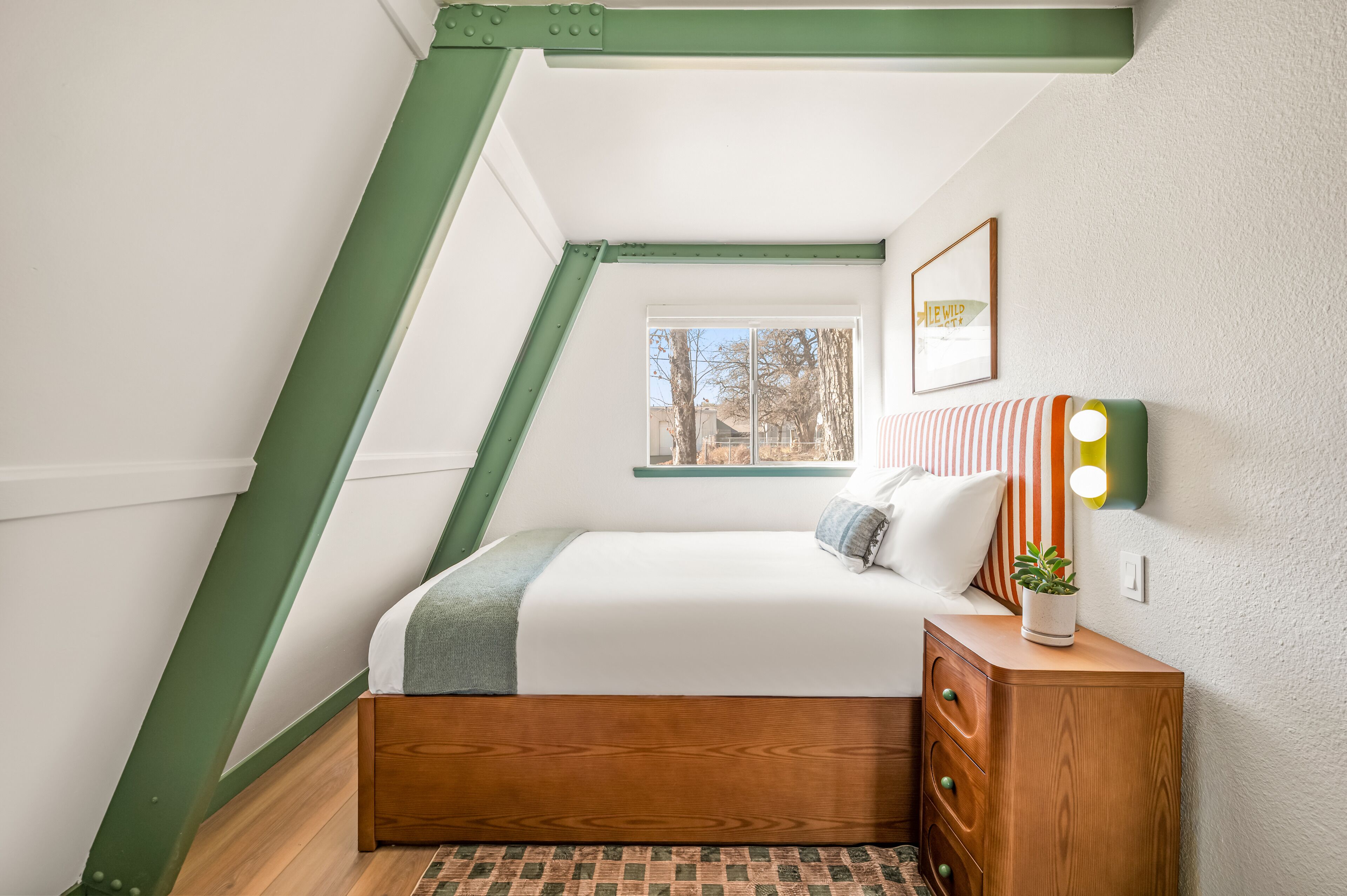 Un dormitorio con techo inclinado, ventana con vistas, cama con almohadas, mesita de noche y una lámpara.