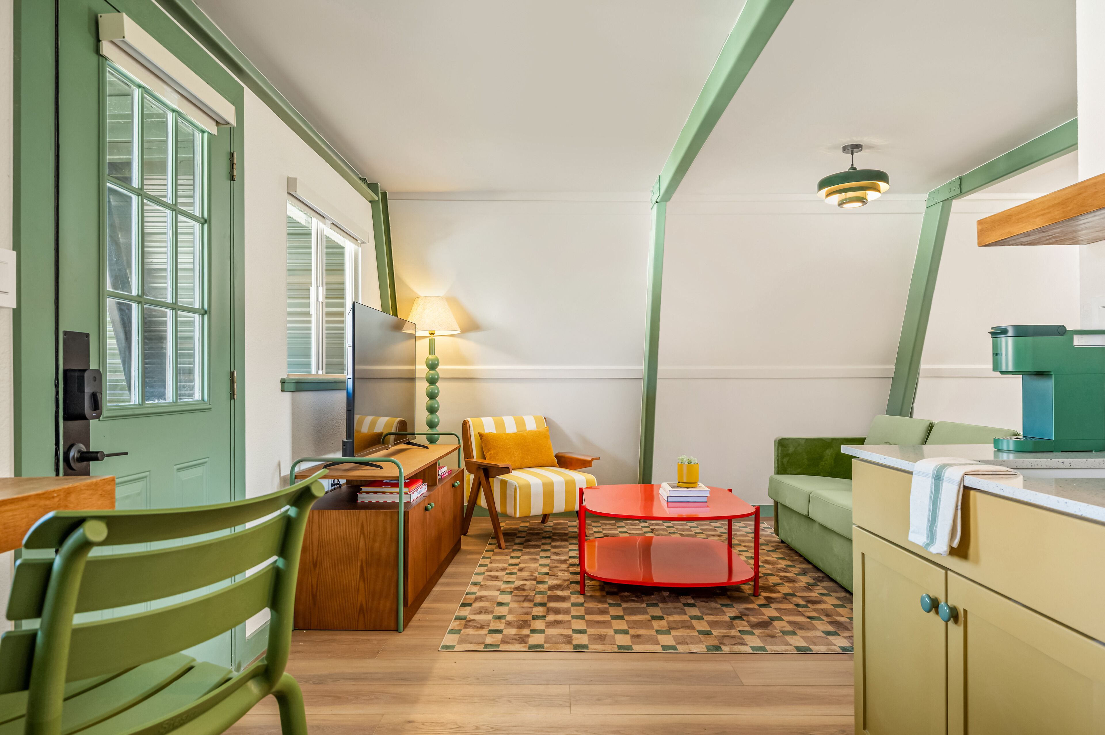 Une pièce compacte et bien éclairée, avec une porte verte, une table en bois, une table basse rouge, un fauteuil à rayures et un canapé vert.
