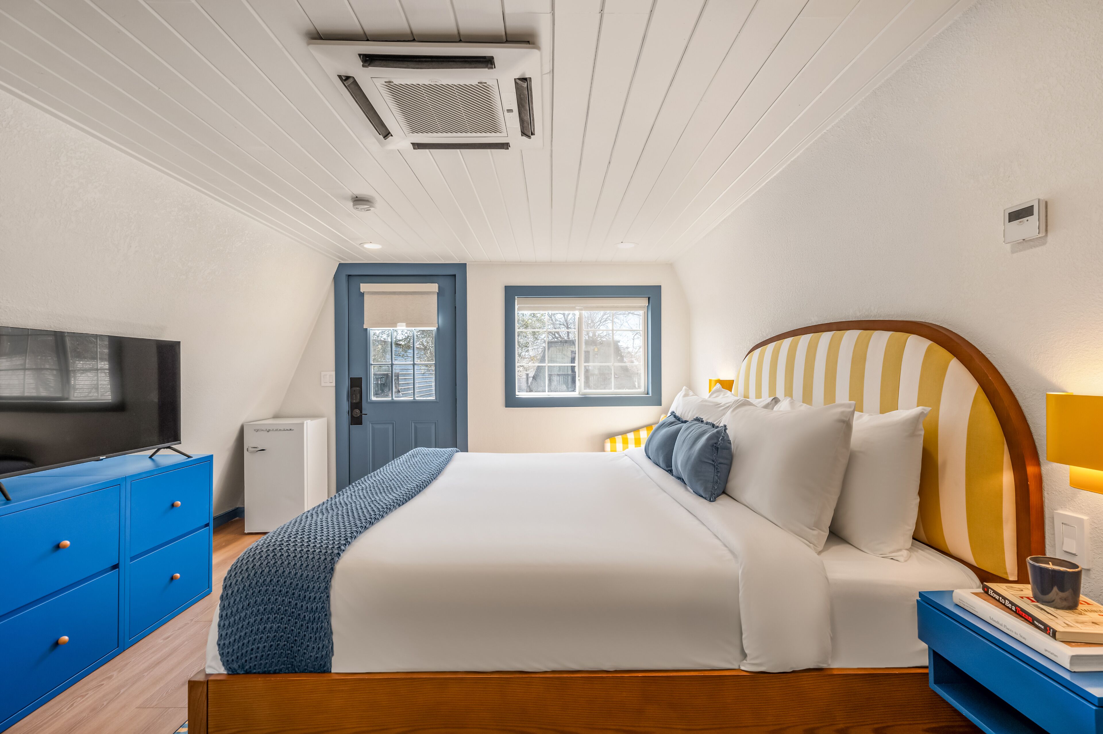 A neatly arranged bedroom with a bed, a TV, a small fridge, and a window.