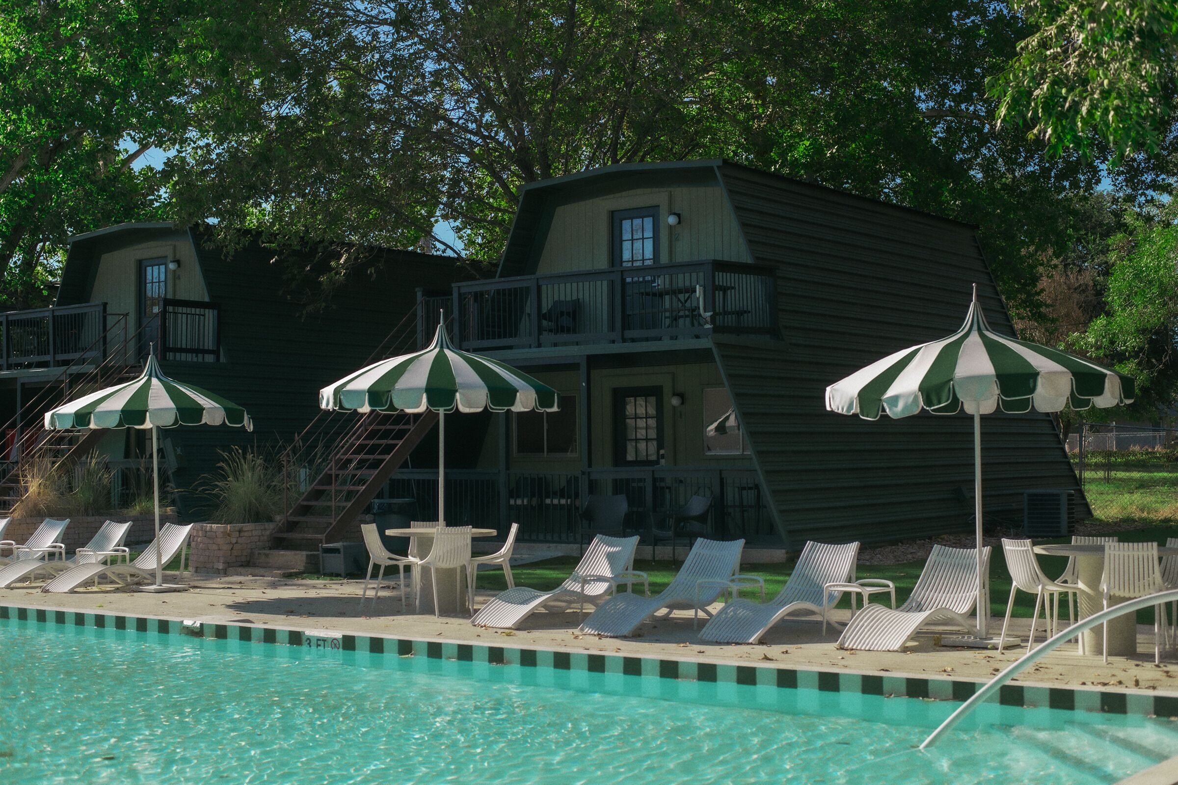 A pool area with lounge chairs and striped umbrellas, a building with a balcony, and a staircase leading to the upper level.
