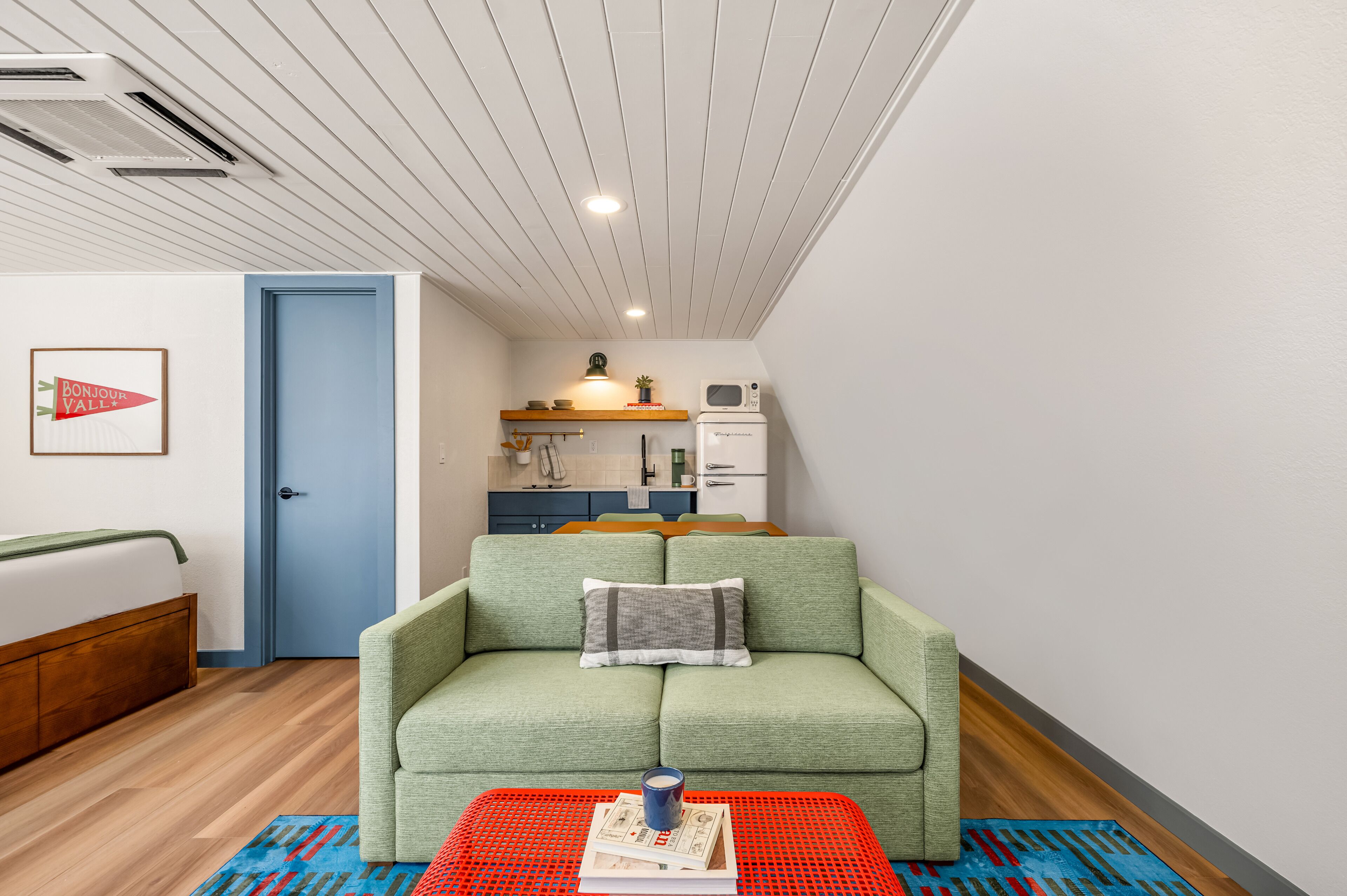 A room with a green sofa, a red table with books, a kitchen area, and a blue door.