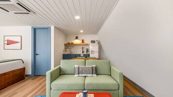 A room with a green sofa, a red table with books, a kitchen area, and a blue door.