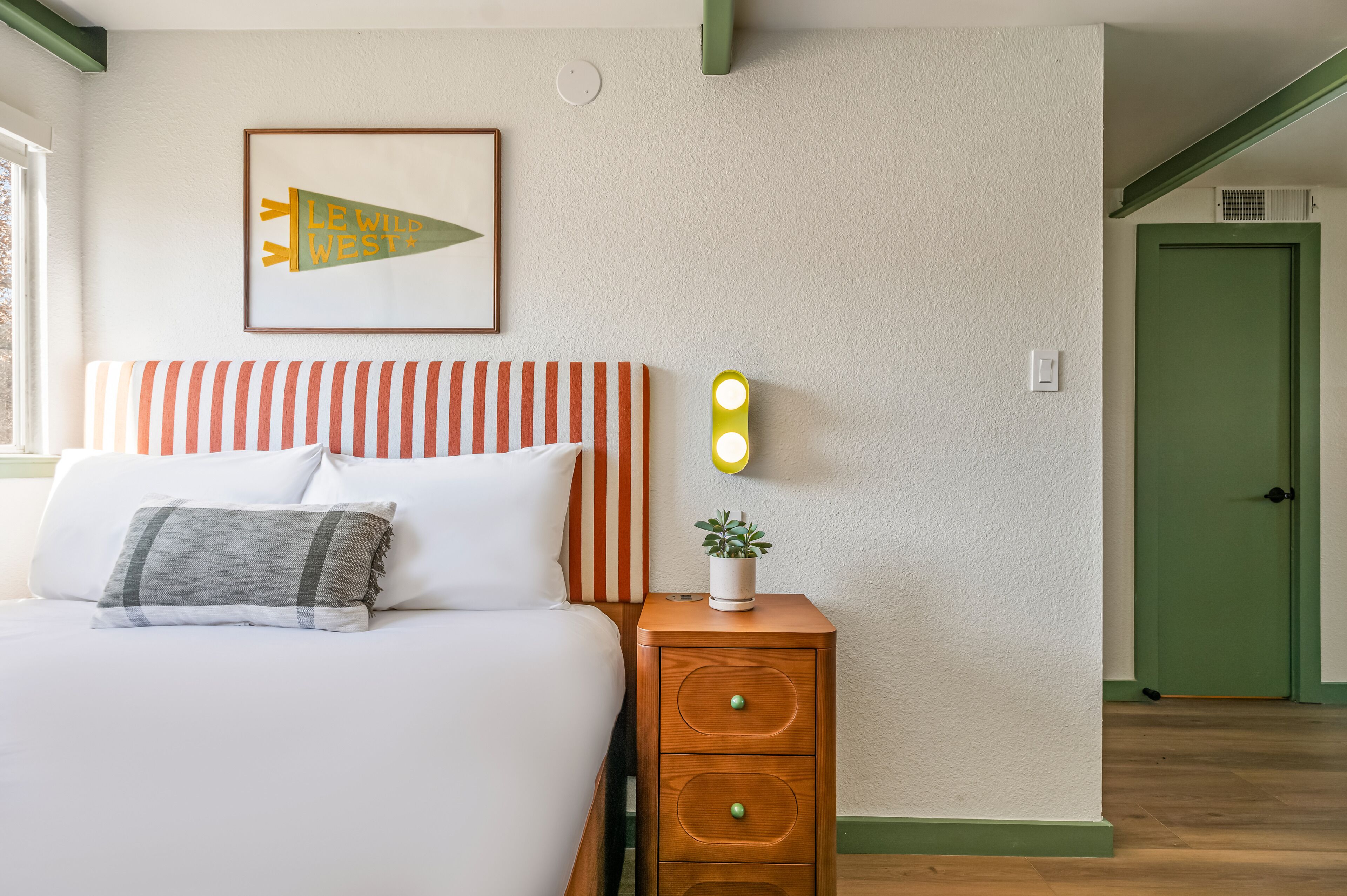 A neatly made bed with striped pillows, a bedside table with a plant, and a framed picture on the wall.