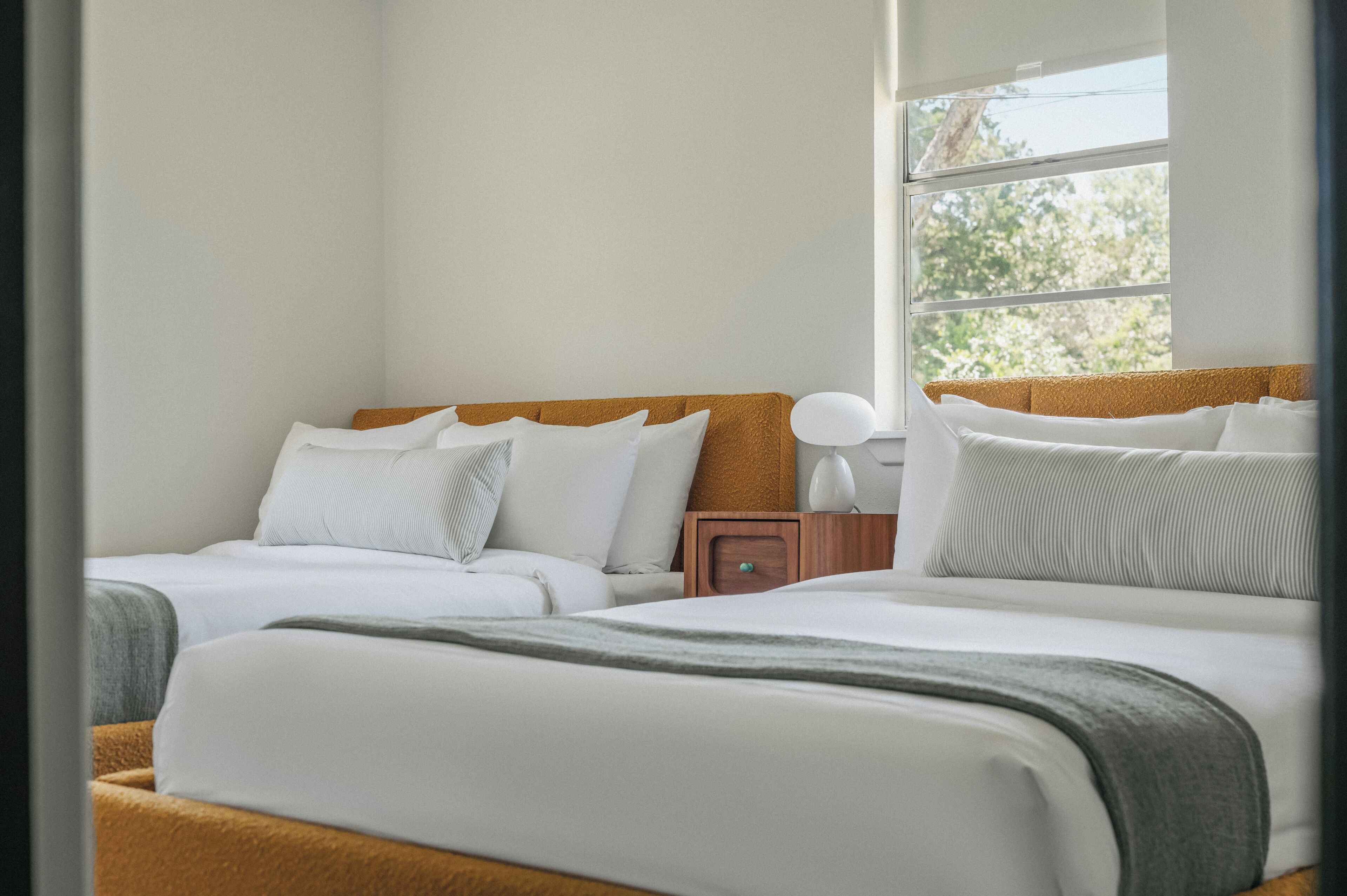 A hotel room with two beds, a nightstand, a lamp, and a window with a view of trees.
